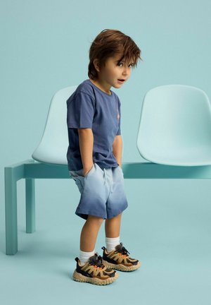 Jonge jongen in blauwe gradient shorts, marineblauw T-shirt en sneakers staat met handen in zakken naast twee lichtblauwe stoelen op een bijpassende achtergrond.