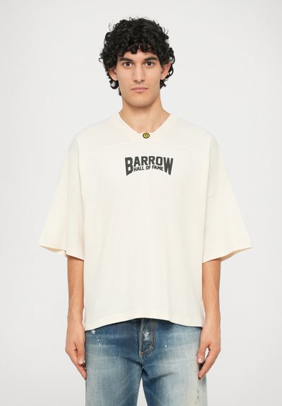 Oversized cream T-shirt with a V-neck, featuring a printed black logo "BARROW HALL OF FAME" across the chest. Worn with distressed blue jeans.