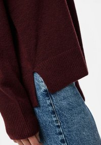 Burgundowy sweter dziany z prążkowanymi mankietami i bocznymi rozcięciami, w połączeniu z jasnoniebieskimi dżinsami z zapięciem na guziki i wyblakłą teksturą.