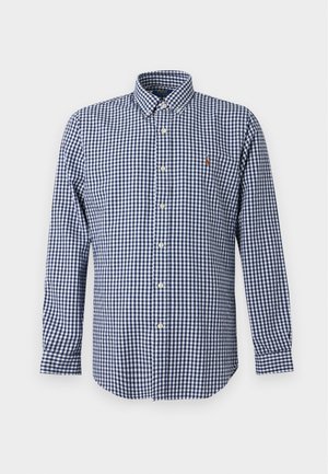 Homme portant une casquette bleu marine, une chemise à carreaux bleue et blanche à manches longues, un pantalon blanc ample et des baskets blanches, debout sur fond blanc.