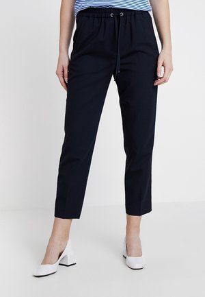 Mujer vistiendo pantalones negros hasta el tobillo con cinturilla elástica y cordón, combinados con zapatos blancos de tacón ancho y una blusa azul a rayas.