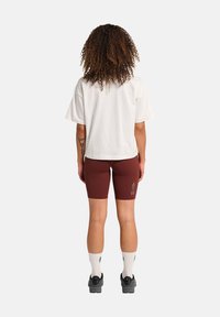 Hvid kortærmet skjorte, bordeaux cykelshorts med grå "ATW" print, hvide ankelstrømper og sorte sko. Naturligt krøllet hår synligt.