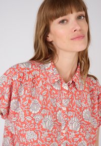 Blusa de manga corta en color coral con un estampado floral en blanco, con cuello clásico y cierre de botones en la parte delantera. Tejido ligero, corte relajado.