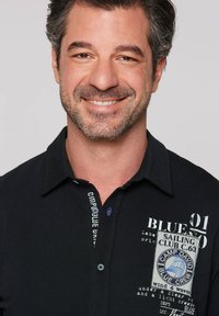 Polo negro con tejido texturizado, que presenta impresiones de logotipos en azul y blanco, botones en el cuello y un detalle de etiqueta cosida.