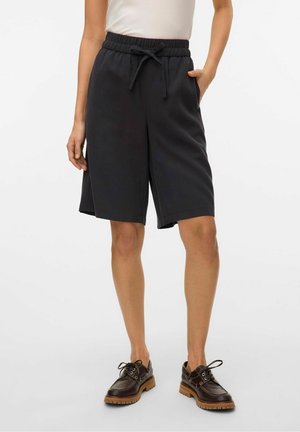 Person trägt schwarze knielange Shorts mit Kordelzug, weißes Oberteil und dunkelbraune Schnürschuhe mit groben Sohlen, steht mit einer Hand in der Tasche.