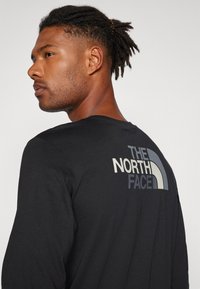 Černá dlouhá tričko s kulatým výstřihem, na zadní straně s logem "THE NORTH FACE" v světle šedé a modré barvě. Hladká textura látky.