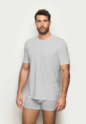 Mann mit kurzen dunklen Haaren und Bart, der ein hellgraues, kurzärmliges T-Shirt und passende Boxershorts trägt, vor schlichtem Hintergrund stehend.