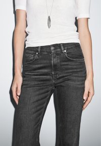 Jean noir en denim avec une coupe ajustée, cinq poches et des coutures visibles. Associé à une chemise blanche à manches courtes et un collier en pendentif argenté.