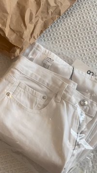 Pantaloni in denim bianco in confezione di plastica con zip laterale. Le caratteristiche includono una vita, tasca sul retro e dettagli con bottoni in tono argento.
