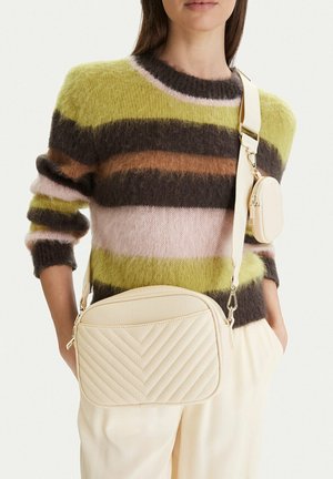 Osoba nosící pruhovaný chlupatý svetr v zelených, hnědých a růžových tónech, nesoucí krémovou prošívanou kabelku přes rameno s malou připevněnou kapsičkou.