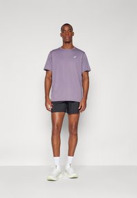 Lilla trænings T-shirt med korte ærmer og logo, parret med sorte shorts, hvide sokker og hvide løbesko med grønne detaljer.