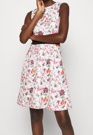 Mujer con vestido blanco sin mangas con estampado floral en rosa, naranja y morado, cinturón negro en la cintura, de pie con un brazo cruzado y la mano cerca de la cara.