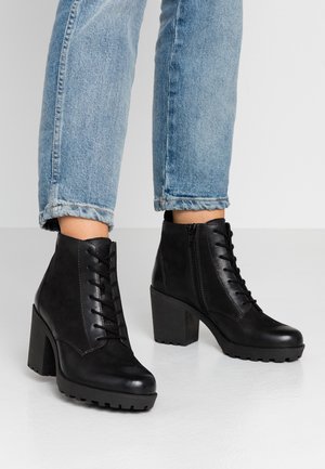Person wearing light blue jeans and black lace-up ankle boots with chunky heels on a plain white floor.