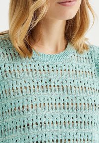 Ljusblå stickad tröja med ett strukturerat mönster och vertikala öppningar. Halsringningen är rund och tyget verkar mjukt och ventilerande.