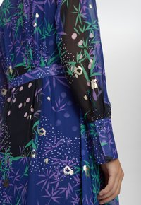 Langsleepsjurk in doorschijnend zwart en marineblauw stof, versierd met groene bamboe en paarse bloemenpatronen, met een strik taille en knoopmanchetten.