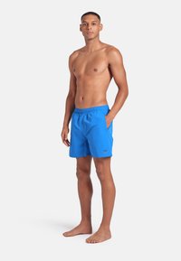 Giovane uomo in piedi a piedi nudi, che indossa pantaloni da bagno blu brillante con cintura elastica e mano nella tasca, sfondo neutro.