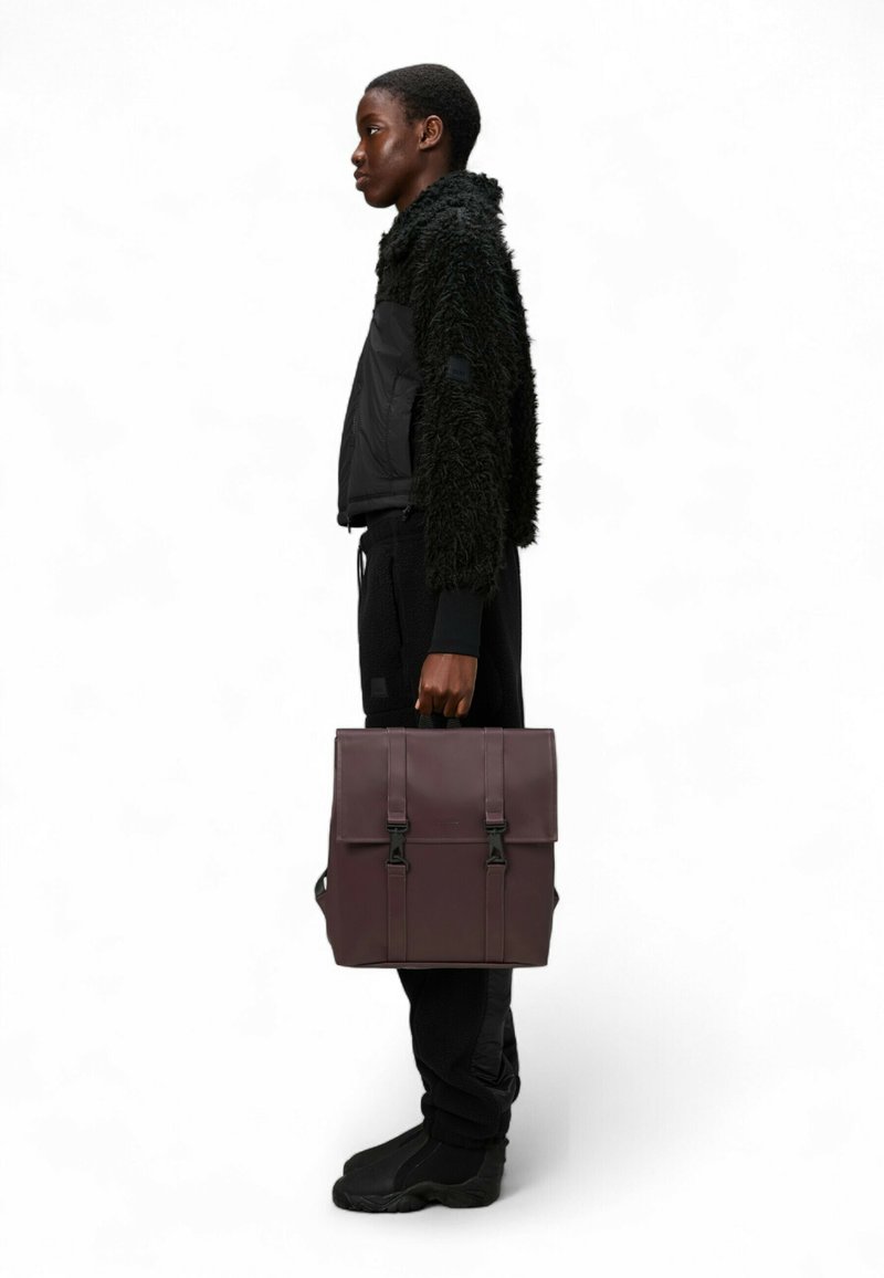 Zaino in pelle bordeaux con doppi fibbie, superficie liscia, forma quadrata. Modello che indossa una giacca nera di pelliccia e pantaloni scuri, vista di profilo.