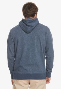 Sudadera con capucha azul de textura suave. Presenta un bolsillo canguro en la parte delantera y puños acanalados en las muñecas y en el dobladillo inferior.