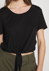 T-shirt noir à manches courtes avec un col rond et un nœud noué à la taille, associé à un pantalon vert olive. Tissu en coton, coupe décontractée.