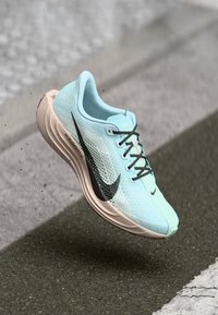 Helle blaue und beigefarbene Nike-Laufschuhe mit schwarzem Swoosh, in der Luft über strukturierter Fahrbahn, wobei Sohle und Schnürsenkel deutlich sichtbar sind.