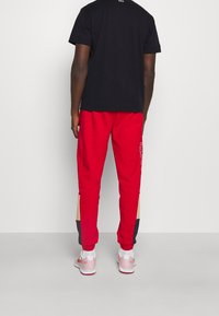 Osoba stojąca tyłem, ubrana w czarną koszulę z krótkim rękawem, czerwone dresy z czarnymi i beżowymi blokami kolorów oraz białe i różowe sneakersy.