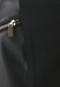 Bolsa preta composta por couro liso e tecido texturizado, com um fecho de correr prateado e um puxador com a marca, realçando o contraste entre os materiais.