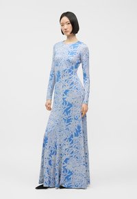 Γυναίκα που στέκεται φορώντας ένα μακρυμάνικο, μακρύ μέχρι το πάτωμα μπλε φόρεμα με λευκό φυλλώδες σχέδιο και μαύρα μυτερά παπούτσια σε απλό φόντο.