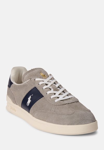 Szürke velúr sneaker, tengerészkék kiegészítőkkel, fehér fűzős kialakítással és arany logóval. A talpa krémszínű és enyhén párnázott.
