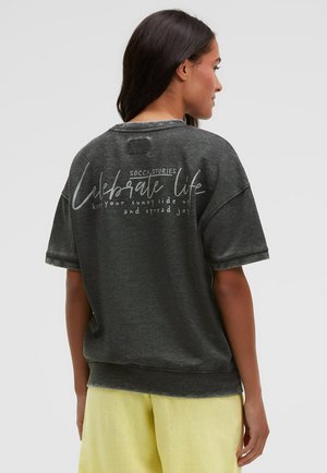 Frau mit dunklen Haaren trägt ein dunkelgraues Hemd mit dem Aufdruck „Celebrate life“ auf der Rückseite und hellgelbe Hosen, steht vor einem schlichten hellen Hintergrund.