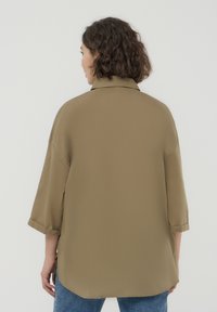 Camicia kaki oversize con colletto, maniche arrotolate e orlo curvo. Il tessuto appare morbido e leggero.