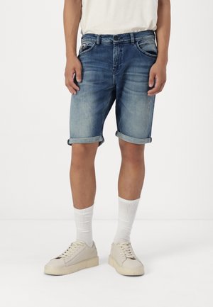 Homme portant un short en denim bleu retroussé, des chaussettes blanches montantes et des baskets couleur crème, debout devant un fond blanc uni.