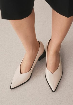 Woman wearing beige pointed-toe low block heel shoes standing on a light-colored floor with black pants.