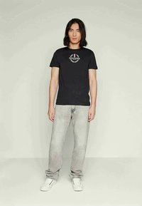 T-shirt de algodão preta apresenta um logotipo com design de louros em branco. Combinada com jeans cinza claro e ténis brancos, apresentada sobre um fundo liso.