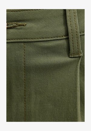 Zbliżenie na oliwkowozieloną tkaninę z pętelką na pasek i widocznym przeszyciem na parze spodni.