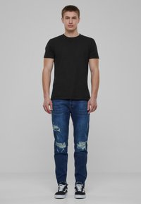 T-shirt noir à manches courtes, jean bleu déchiré avec des accents de déchirure, porté avec des baskets noires et blanches, sur fond uni.