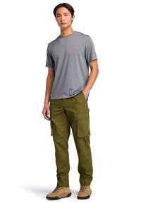 T-shirt grigio a maniche corte con logo arancione, abbinato a pantaloni cargo verde oliva con tasche laterali. Modello che indossa stivali da escursionismo beige.
