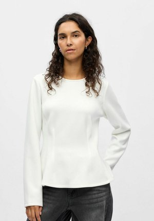 Weißes langärmeliges Oberteil mit rundem Halsauschnitt und strukturierter Form. Glattes Gewebe mit dezenten Nähten, kombiniert mit dunkelgrauen Jeans.