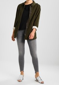 Giacca verde oliva con chiusura zip, camicia nera sotto, jeans skinny grigi e tacchi aperti argento. Maniche arrotolate e dettagli sulle spalle.