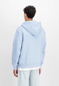 Világoskék pulóver kapucnis felső, puha, texturált anyagból, tágas kapucnival és bordás mandzsettákkal, fehér, rétegezett blúz fölé húzva.