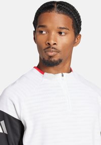 Maglia sportiva bianca a mezza zip con collo rosso e maniche nere. Presenta un tessuto testurizzato con strisce orizzontali e una zip al collo.