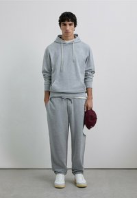 Sudadera gris con capucha y cordones, combinada con pantalones deportivos grises a juego. Zapatillas blancas y gorra burdeos sostenida en la mano.