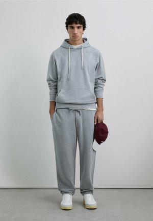 Sudadera gris con capucha y cordones, combinada con pantalones deportivos grises a juego. Zapatillas blancas y gorra burdeos sostenida en la mano.