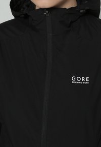 Giacca da corsa nera con zip e cappuccio, con logo bianco "GORE RUNNING WEAR" sul petto a sinistra.