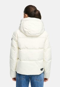 Donna con capelli castani raccolti in una coda di cavallo, che indossa una giacca a vento bianca con cappuccio e jeans blu, di spalle su uno sfondo semplice.