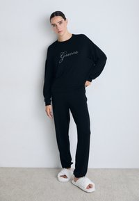 Frau in schwarzem Guess Sweatshirt und Hose mit weißen, flauschigen Hausschuhen, die gegen eine schlichte helle Wand und einen Teppichboden steht.