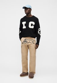 Μαύρο πλεκτό πουλόβερ με λευκά γράμματα "IC", καφέ παντελόνι cargo με σχέδιο τρέχοντος γάτου και καφέ παπούτσια slip-on με λουλουδένιο κέντημα.