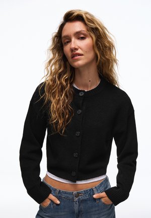 Femme aux longs cheveux ondulés porte un cardigan noir boutonné sur un haut blanc, jean bleu, debout les mains dans les poches sur fond blanc.