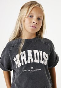 Grijze sweatshirt met een ronde halslijn, voorzien van een grote witte opdruk "PARADISE" en kleinere tekst eronder. De stof heeft een licht versleten textuur.