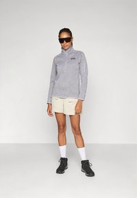 Szürke polárpulóver magas nyakkal és félcipzárral, bézs rövidnadrággal párosítva. Fekete túrabakancs és fehér zokni egészíti ki a ruházatot.