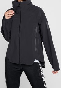 Personne portant une veste noire à col montant avec fermeture éclair et un pantalon noir texturé avec des bandes blanches sur les côtés, main tenant la fermeture éclair de la veste.